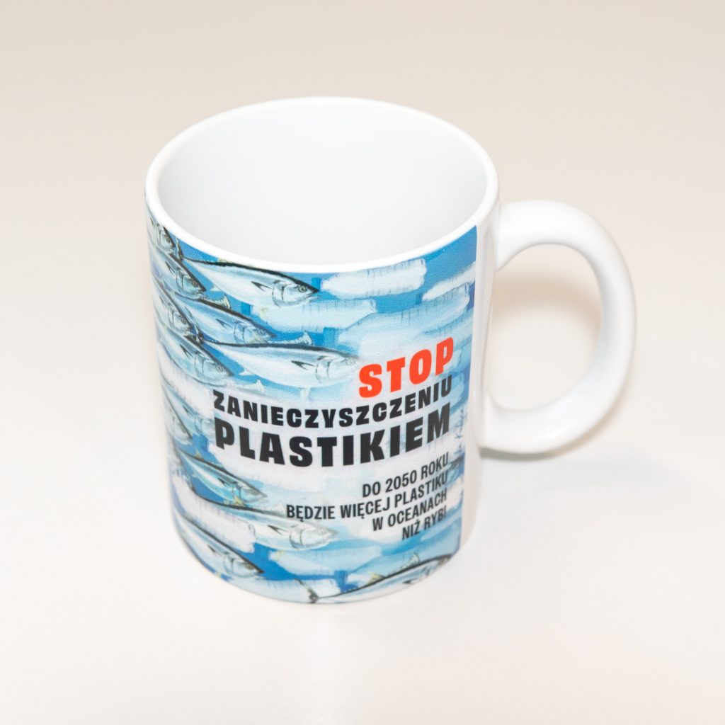 Biały kubek z nadrukiem „Stop zanieczyszczeniu plastikiem” i grafiką ryb pływających wśród plastikowych odpadów.