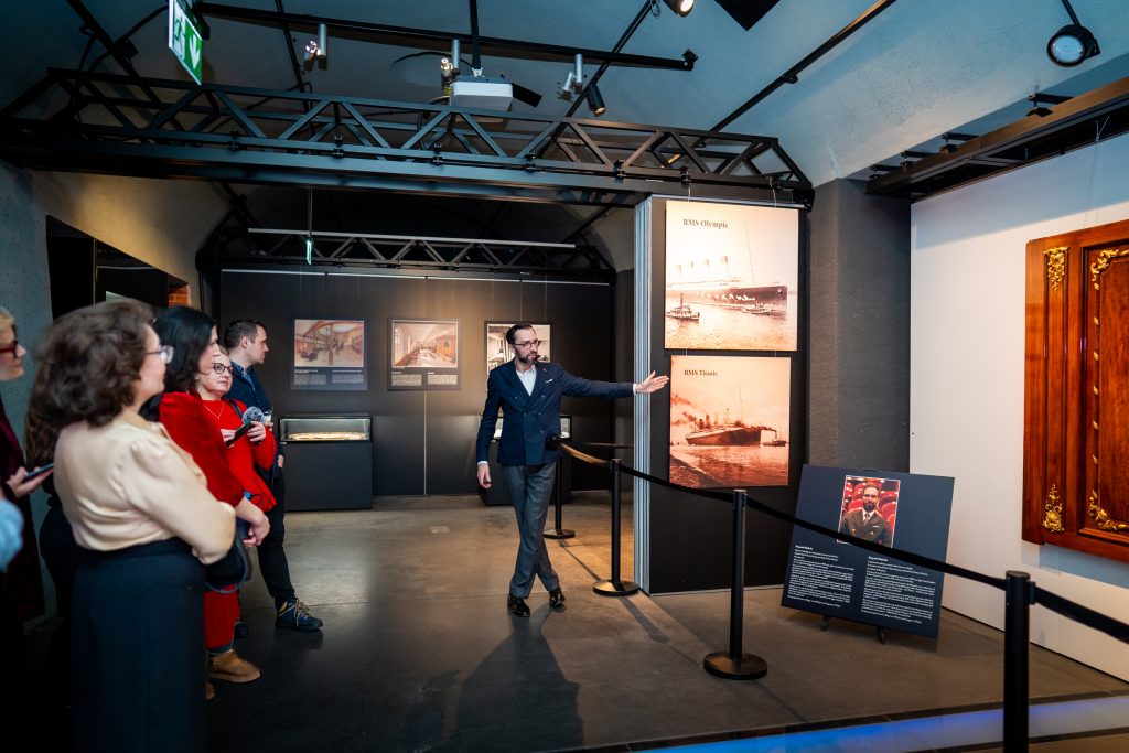 Kurator prezentuje fragment wystawy poświęconej liniowcom Titanic, Britannic i Olympic, wskazując na panel boazeryjny, podczas gdy grupa zwiedzających uważnie słucha