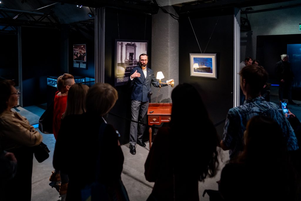 Kurator prezentujący element wystawy przed grupą odwiedzających, na tle fotografii i artefaktów z epoki wielkich liniowców oceanicznych.