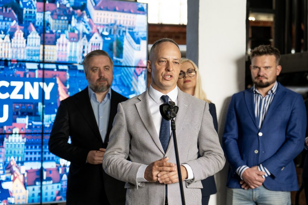 Mężczyzna wypowiada się do mikrofonu. W tle inni prelegenci