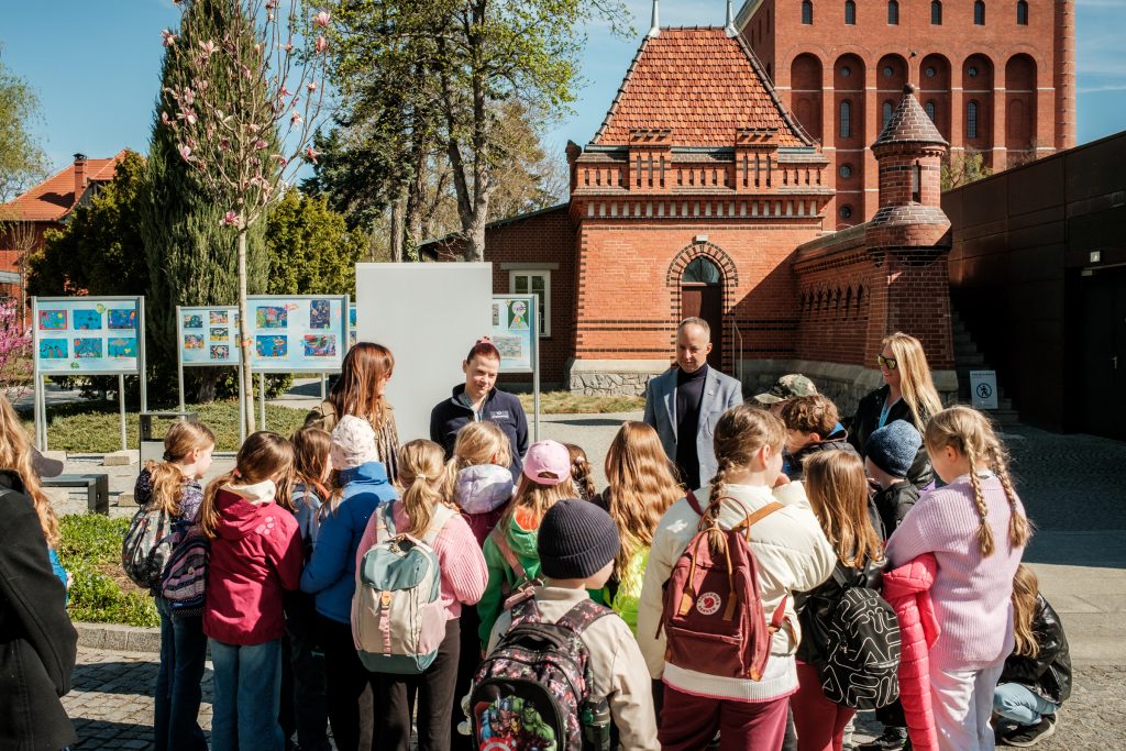 Na zdjęciu dzieci w towarzystwie dorosłych na dworze. W tle zabytkowa architektura. Dzieci z zainteresowaniem słuchają dorosłych.