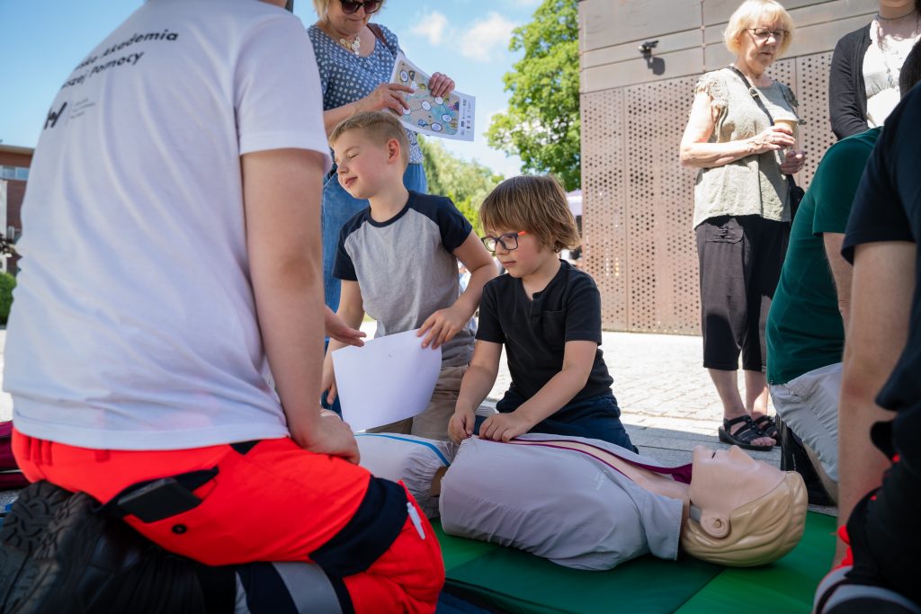Dzieci ćwiczą udzielanie pierwszej pomocy na fantomie pod okiem instruktora podczas warsztatów edukacyjnych na świeżym powietrzu