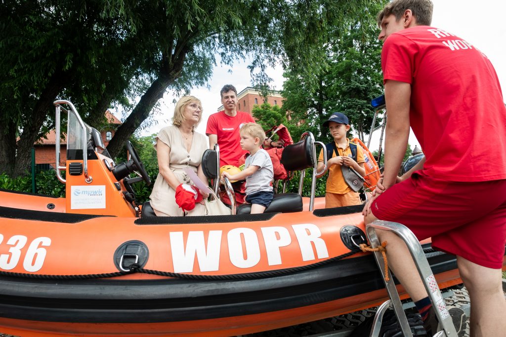 Uczestnicy wydarzenia siedzą w łodzi ratowniczej WOPR, a ratownik prezentuje sprzęt i prowadzi rozmowę edukacyjną z dziećmi i dorosłymi