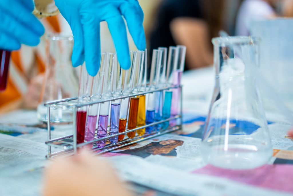 Niebieskie rękawice ochronne nad stojakiem z probówkami wypełnionymi kolorowymi cieczami, ustawionymi na stole laboratoryjnym.