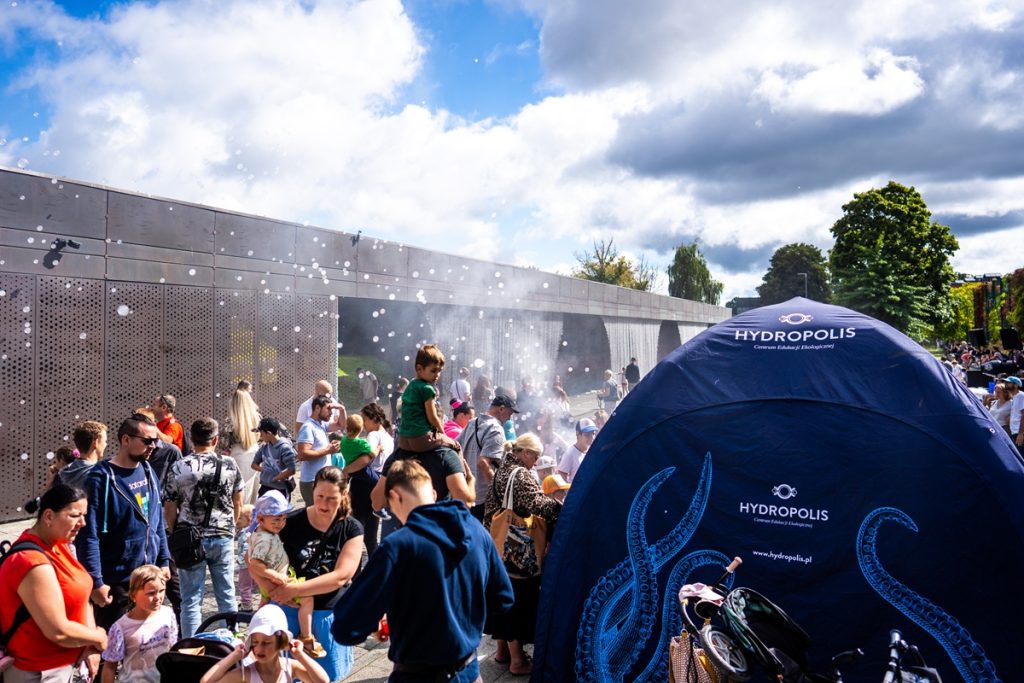 Uczestnicy rodzinnego pikniku przed Hydropolis we Wrocławiu. Na pierwszym planie namiot Hydropolis, w tle wodna mgiełka i odwiedzający korzystający z atrakcji.