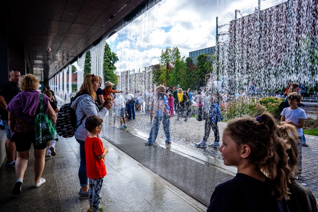 Goście rodzinnego pikniku spacerują przy wodnej kurtynie przed budynkiem Hydropolis.
